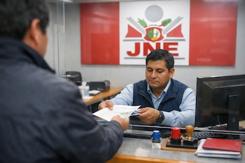 Un empleado del JNE con camisa azul recibe documentos de un ciudadano en un mostrador de atención. El logo del JNE, con el escudo peruano, se ve en la pared detrás del empleado.