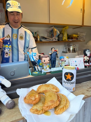 Hombre con camiseta de Argentina y gorra sonríe mientras sostiene una botella, junto a un plato de Khuushuur fritos, una cerveza Sapporo y figuras de fútbol
