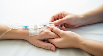 Dos manos entrelazadas sobre una sábana blanca; una mano con catéter intravenoso y adhesivo médico, la otra mano la sujeta.