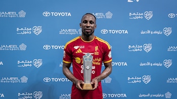 Un hombre con camiseta deportiva roja y amarilla sostiene un trofeo plateado frente a un fondo azul con logotipos.