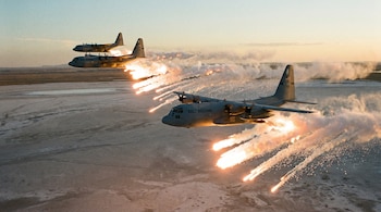 Tres aviones C-130 grises vuelan bajo sobre un paisaje árido y plano, liberando estelas de bengalas naranjas y blancas que iluminan el cielo al atardecer.