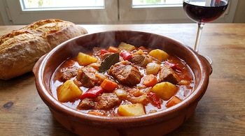 Cazuela de barro con estofado de carne de cerdo, patatas, zanahorias, pimientos, cebolla y hoja de laurel. Barra de pan y copa de vino tinto en mesa de madera.
