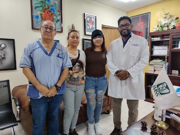 Cuatro personas de pie en un consultorio; dos hombres en vestimenta médica, una mujer con gorra y vendaje nasal, y otra mujer. Bandera del IMSS