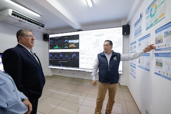 Dos hombres en una sala: uno de traje y gafas mira a la derecha, otro con chaleco señala pósteres. Detrás, una gran pantalla muestra gráficos y datos