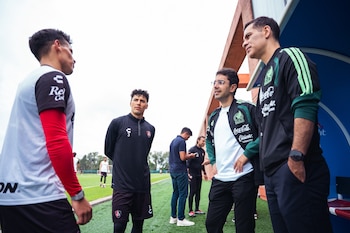 Cuatro hombres conversan al borde de una cancha de fútbol en un día despejado, dos de ellos con ropa deportiva