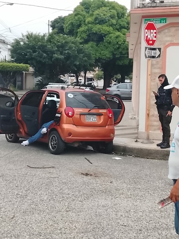 Piernas de una persona sobresalen de un Chevrolet Spark naranja con puertas abiertas en una calle; un agente de policía uniformado observa la escena cerca de un poste con señal de "PARE"