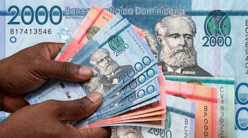 Unas manos de piel morena sostienen un abanico de billetes de dos mil pesos dominicanos sobre un fondo con un billete de mayor tamaño.