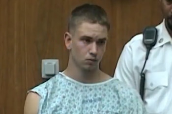 Un hombre joven con bata médica estampada permanece de pie junto a un agente de policía con camisa blanca frente a una pared de madera