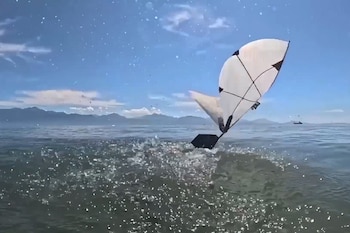 Vehículo blanco con vela negra emerge del agua. Grandes salpicaduras. Cielo azul con nubes, montañas en el horizonte, otro objeto flotando en el agua lejana