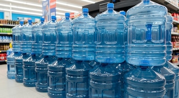 Hileras de botellones de agua grandes de plástico azul transparente apilados en un pasillo de supermercado, con estanterías al fondo.