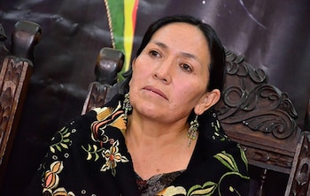 Primer plano de Tomasa Yarhui, una mujer con cabello oscuro y aretes de cuentas, vistiendo una prenda negra bordada con flores, sentada en una silla de madera tallada
