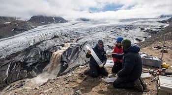 Tres científicos con ropa de invierno examinan un núcleo de hielo transparente en un glaciar gris con grietas, agua marrón y montañas al fondo con nubes.