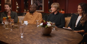 Vista lateral de cuatro personas, incluyendo a Owen Wilson y Glen Powell, sentadas alrededor de una mesa de madera oscura con bebidas y un centro floral