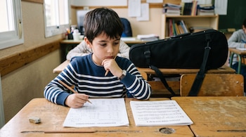 Un niño sentado frente a un escritorio con hojas de matemáticas y una partitura de violín, sosteniendo un lápiz junto al rostro.