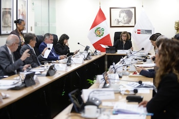 Autoridades sentadas alrededor de una mesa de reuniones con micrófonos, documentos y la bandera peruana al fondo