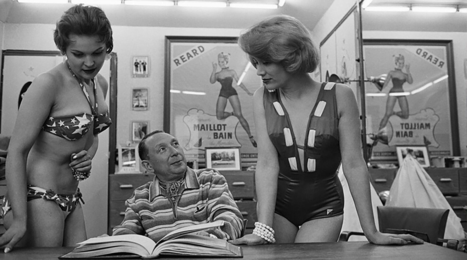 Louis Réard, creador del bikini, junto a dos modelos en su taller de diseño en París, durante la década de 1950. En las paredes, afiches promocionales de sus trajes de baño con el nombre de la piscina Molitor, donde presentó por primera vez su creación en 1946 (AFP)