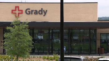 Primer plano del exterior del edificio Grady con el logo rojo de una cruz y el nombre, ventanas de vidrio, un árbol frondoso y arbustos delante