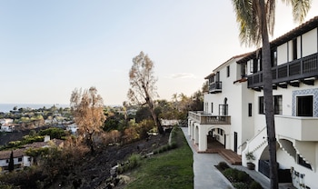 Fachada de una casa blanca de estilo español con balcones oscuros, junto a una pendiente con árboles quemados y palmeras