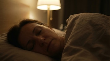 Primer plano de una mujer de 50 años durmiendo de lado en la cama, con el rostro sudoroso y los ojos cerrados. Una lámpara tenue ilumina la habitación oscura.