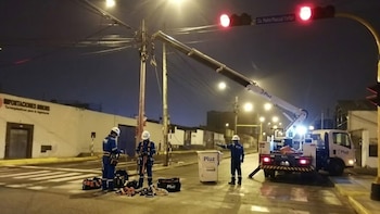 Tres trabajadores con uniformes azules y cascos blancos operan junto a un camión elevador de Pluz cerca de un poste de luz y un semáforo en rojo en la calle