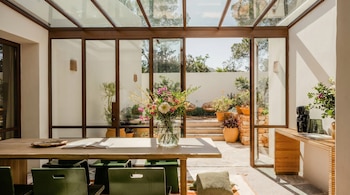 Un luminoso comedor con techo y paredes de cristal que dan a un patio con escaleras y plantas. Una mesa de madera con sillas verdes y un jarrón de flores en el centro.