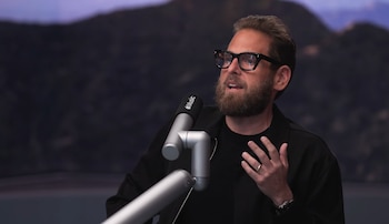 Un hombre barbudo con gafas de montura negra, Jonah Hill, gesticula con la mano mientras habla por un micrófono de Apple Music