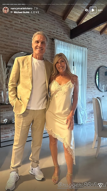 Hombre y mujer sonrientes posan abrazados. Él con traje beige y zapatillas; ella con vestido crema de encaje y tacones. Fondo con pared de ladrillo y techo de madera