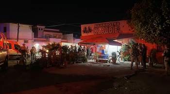 Vista nocturna de un local de abarrotes con toldos naranjas, varias personas y un vehículo de emergencia con luces rojas en una calle oscura.