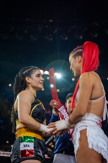 Vista de cerca de dos boxeadoras, Flor Vigna con trenzas y Alana Flores con cabello rojo vibrante, dándose la mano en un ring