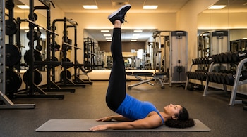 Una mujer con leggings negros y top azul, acostada en una colchoneta gris en un gimnasio, eleva sus piernas estiradas. Se ven máquinas de ejercicio y espejos.