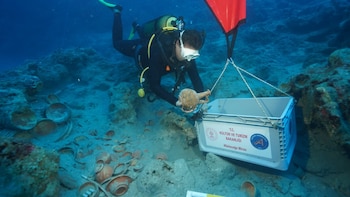 Una persona con equipo de buceo guarda una vasija antigua en una caja blanca rodeada de platos cerámicos en el lecho marino