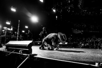 Fotografía monocromática de un músico con sombrero vaquero arrodillado en un escenario iluminado, con otros músicos y el público en el fondo