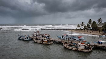 Varios barcos atracados en un muelle, mar con olas grandes, cielo gris con nubes densas, playa con arena y palmeras agitadas por el viento.