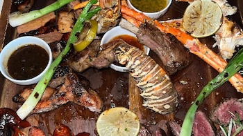 Plato con langosta, patas de cangrejo, trozos de carne asada, limones a la parrilla, cebollas verdes asadas y varios recipientes con salsas
