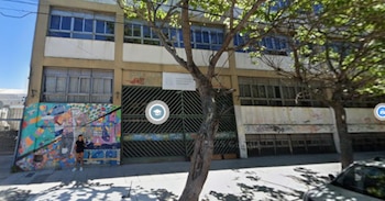 Fachada de un edificio escolar beige de varios pisos con ventanas, una reja metálica y un mural colorido. Un árbol y una persona en la acera