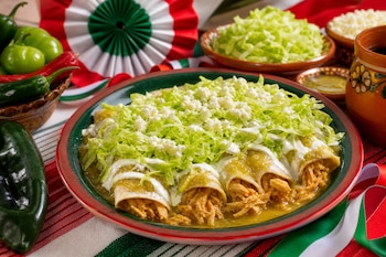 Un plato con enchiladas verdes de pollo cubiertas de lechuga y queso, rodeado de chiles frescos y adorno tricolor.