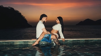 Hombre y mujer sentados al borde de una piscina con agua. Fondo de atardecer, mar y siluetas de montañas. El hombre tiene tatuajes, la mujer cabello largo.