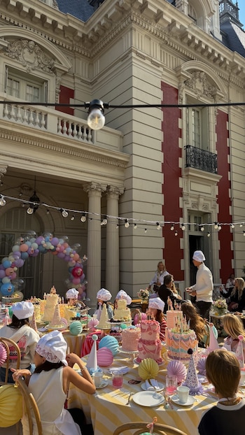 Niñas con gorros de cocinero sentadas a una mesa con tortas decoradas y adornos festivos frente a la fachada de un palacio
