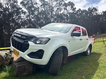 Camioneta pickup Isuzu D-Max blanca estacionada sobre grandes rocas en un campo de hierba verde, con un fondo de árboles y cielo nublado