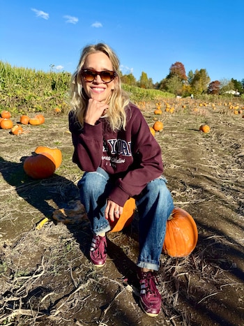 Una mujer con gafas de sol, sudadera granate y pantalones vaqueros sonríe sentada sobre una calabaza en un campo abierto lleno de calabazas