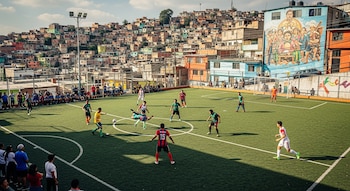 Vista aérea da partida de futebol de 7 no campo artificial de Iztapalapa. Participam atores e espectadores, com prédios e murais ao fundo.