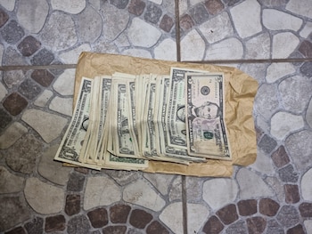 Pila de billetes de uno y cinco dólares estadounidenses sobre un trozo de papel marrón arrugado, colocado en un suelo de baldosas con patrón irregular