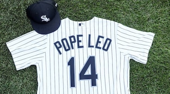 Una camiseta de béisbol blanca a rayas negras con el nombre 'POPE LEO' y el número '14', y una gorra negra con el logo 'Sox', yacen sobre césped verde