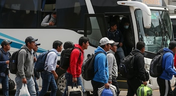 Un grupo de hombres jóvenes con mochilas y gorras camina junto a un autobús interprovincial blanco y azul, algunos subiendo a bordo.