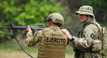 Dos soldados en uniformes de camuflaje. Uno, de espaldas, apunta con un rifle y lleva un chaleco con la palabra "EJÉRCITO". El otro, a su lado, lo guía con una mano en su hombro. El fondo es un bosque verde borroso