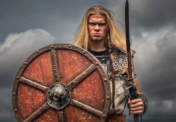 Erling Haaland, con cabello largo y rubio, posa como vikingo sosteniendo un escudo redondo de madera y una espada, bajo un cielo nublado