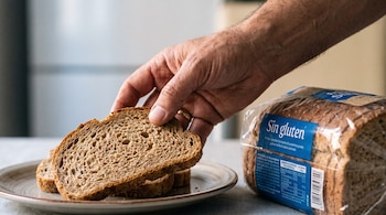 Mano adulta toma una rebanada de pan de trigo de un plato, junto a un pan sin gluten empaquetado con etiqueta azul 'Sin gluten' visible.