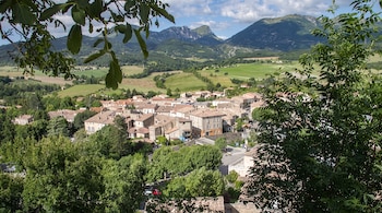 Pueblo con casas de tejas naranjas en un valle, campos verdes, montañas al fondo, árboles frondosos en primer plano, cielo con nubes.