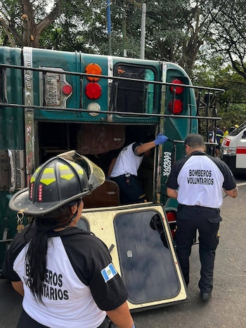 Vista de tres miembros de los Bomberos Voluntarios de Guatemala trabajando alrededor de la parte trasera abierta de un autobús verde en una calle arbolada