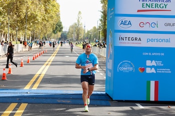 Un hombre con camiseta azul, pantalones cortos oscuros y gafas de sol rojas corre sobre una alfombra azul en la meta de un maratón, con conos naranjas y un cartel de patrocinadores